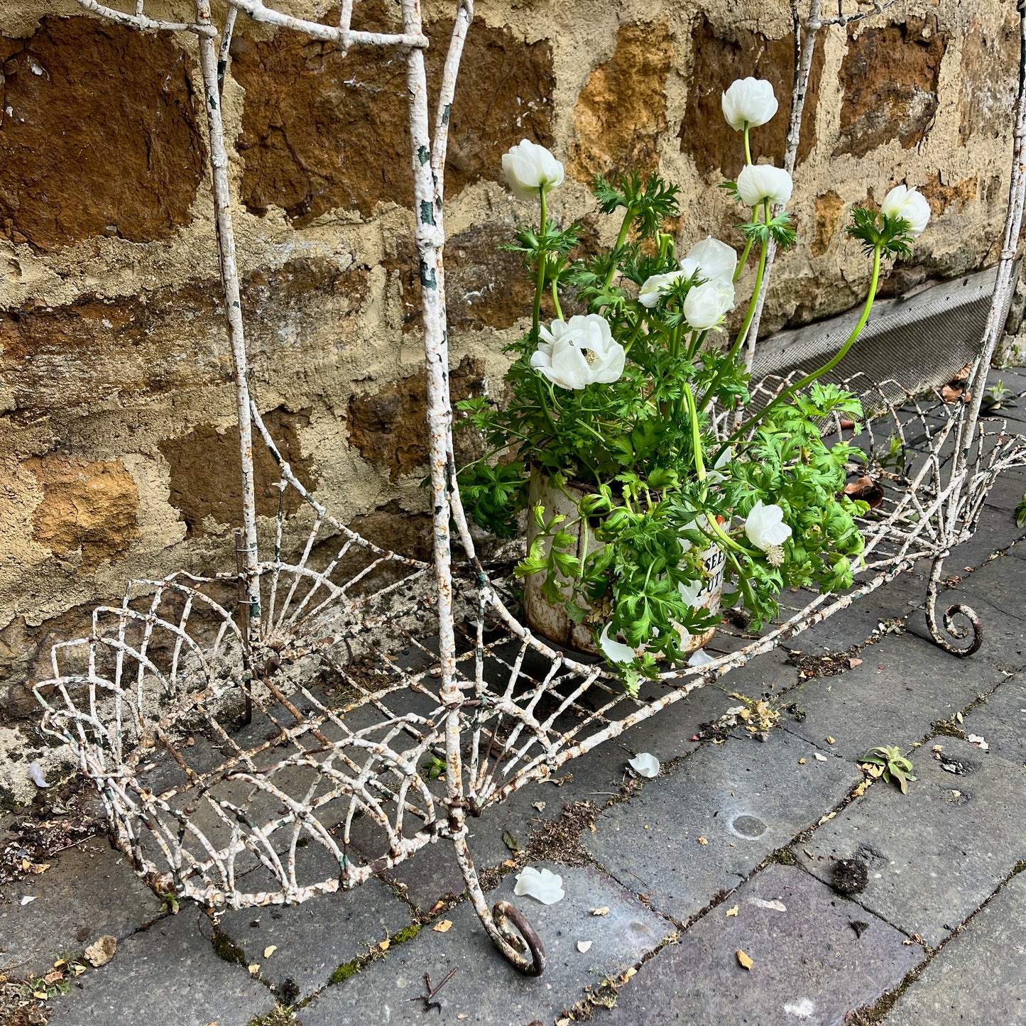 A Victorian decorative plant stand crafted from ornate wrought iron wire, featuring intricate scrollwork and floral patterns. The two-tiered stand, painted in a distressed white finish, supports small potted plants, exuding elegant vintage charm