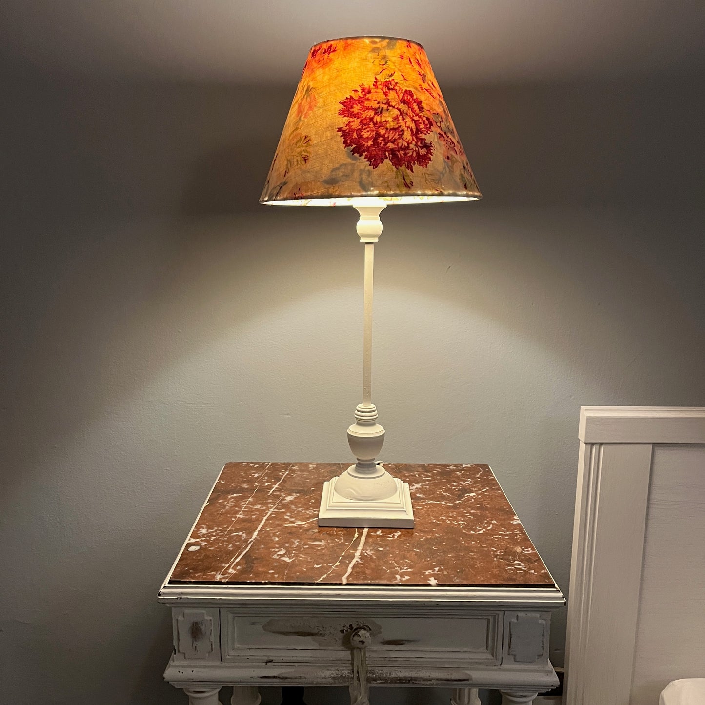 A handmade lampshade made from vintage French linen which sits atop a simple white base. The white base provides a clean, minimalist contrast, highlighting the delicate details of the shade. The overall design evokes a sense of understated sophistication and vintage charm.