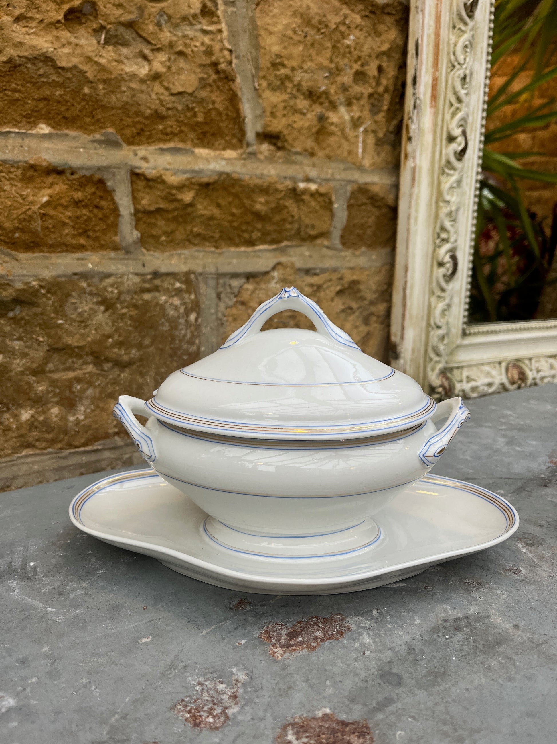 A vintage French sauce boat made of white porcelain with delicate blue and gold   patterns along the edges. The boat has an elegant lid with handle and sits on a matching oval saucer.