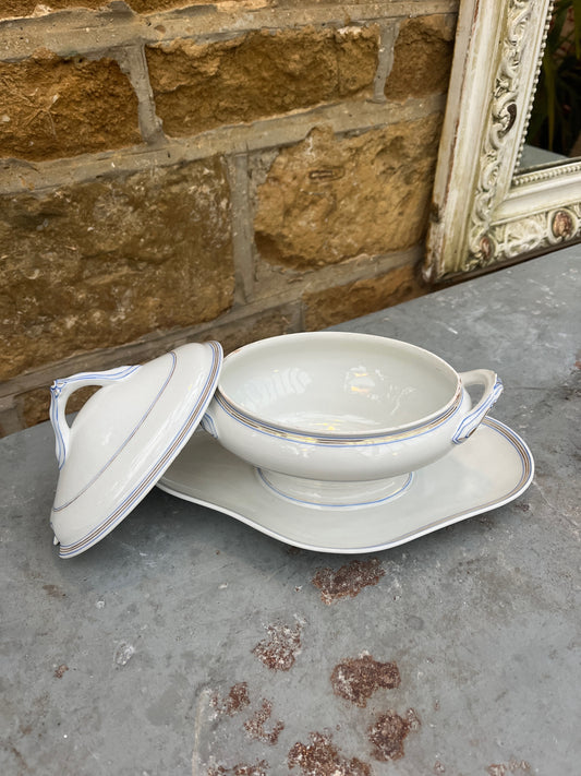 A vintage French sauce boat made of white porcelain with delicate blue and gold   patterns along the edges. The boat has an elegant lid with handle and sits on a matching oval saucer.