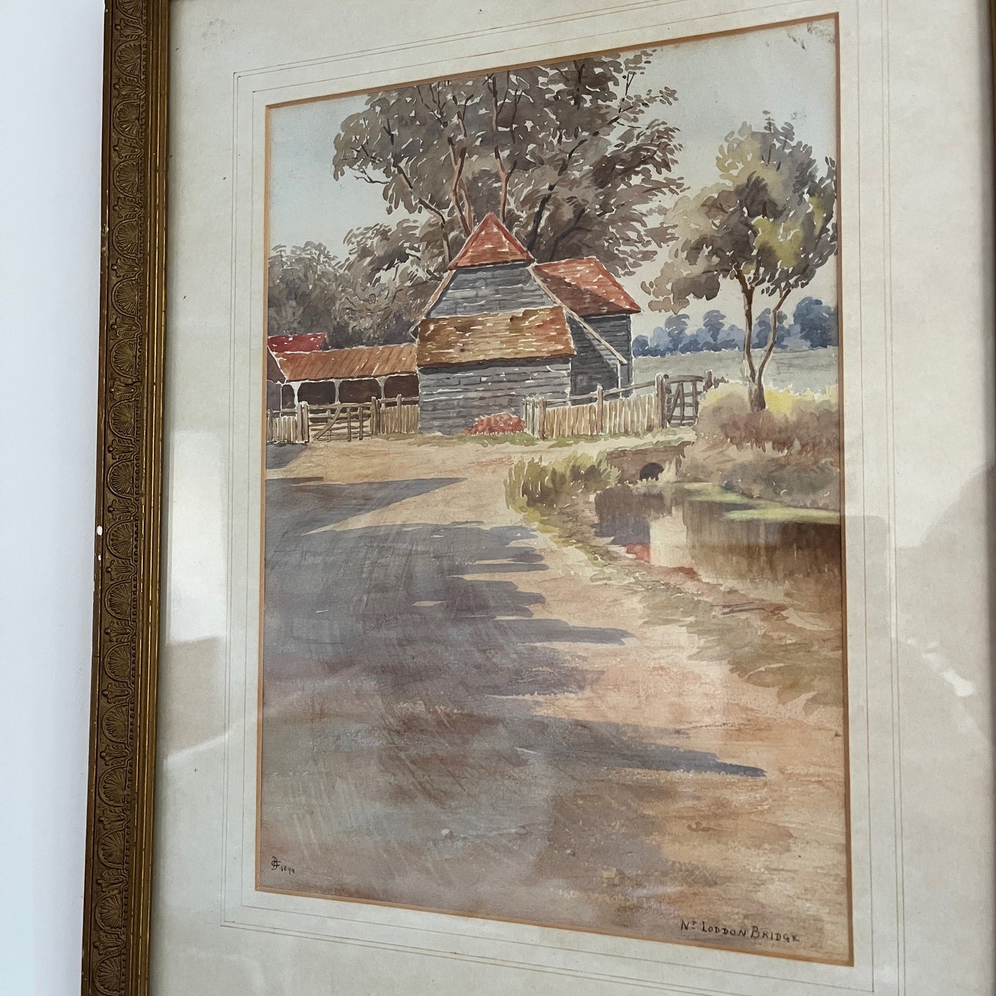 An antique watercolor painting of Loddon Bridge, depicting a serene riverside scene with a stone bridge arching over calm waters. The bridge features weathered stonework, flanked by lush greenery and trees in soft shades of green and brown.
Beside the bridge is an old farm building with a terracotta roof. The overall style is detailed yet gentle, characteristic of traditional watercolor techniques.
