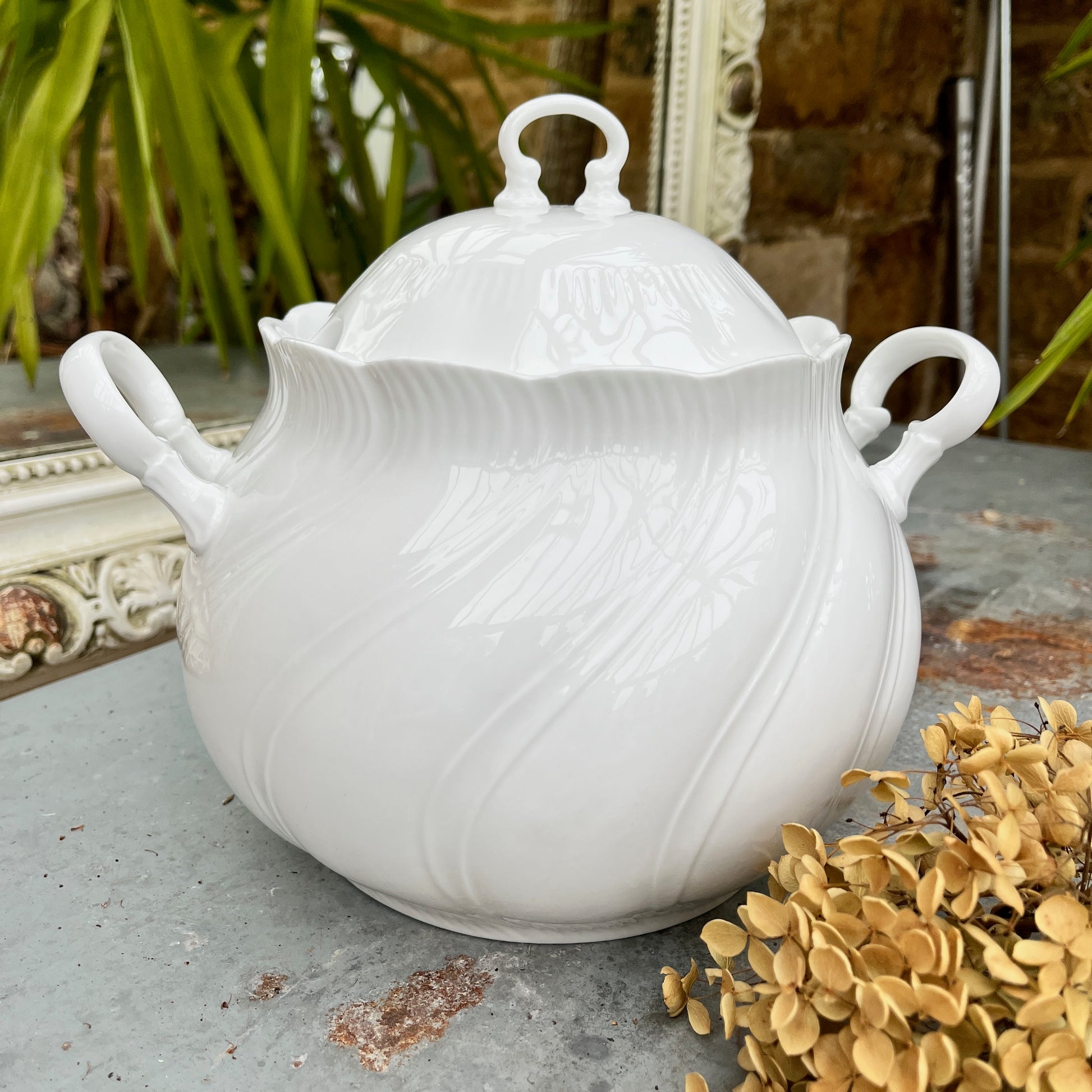 A round, white French-style casserole dish made of glazed ceramic, featuring a smooth, glossy finish. The dish has two small, curved handles on opposite sides and a fitted lid with a simple knob, exuding elegant, timeless charm