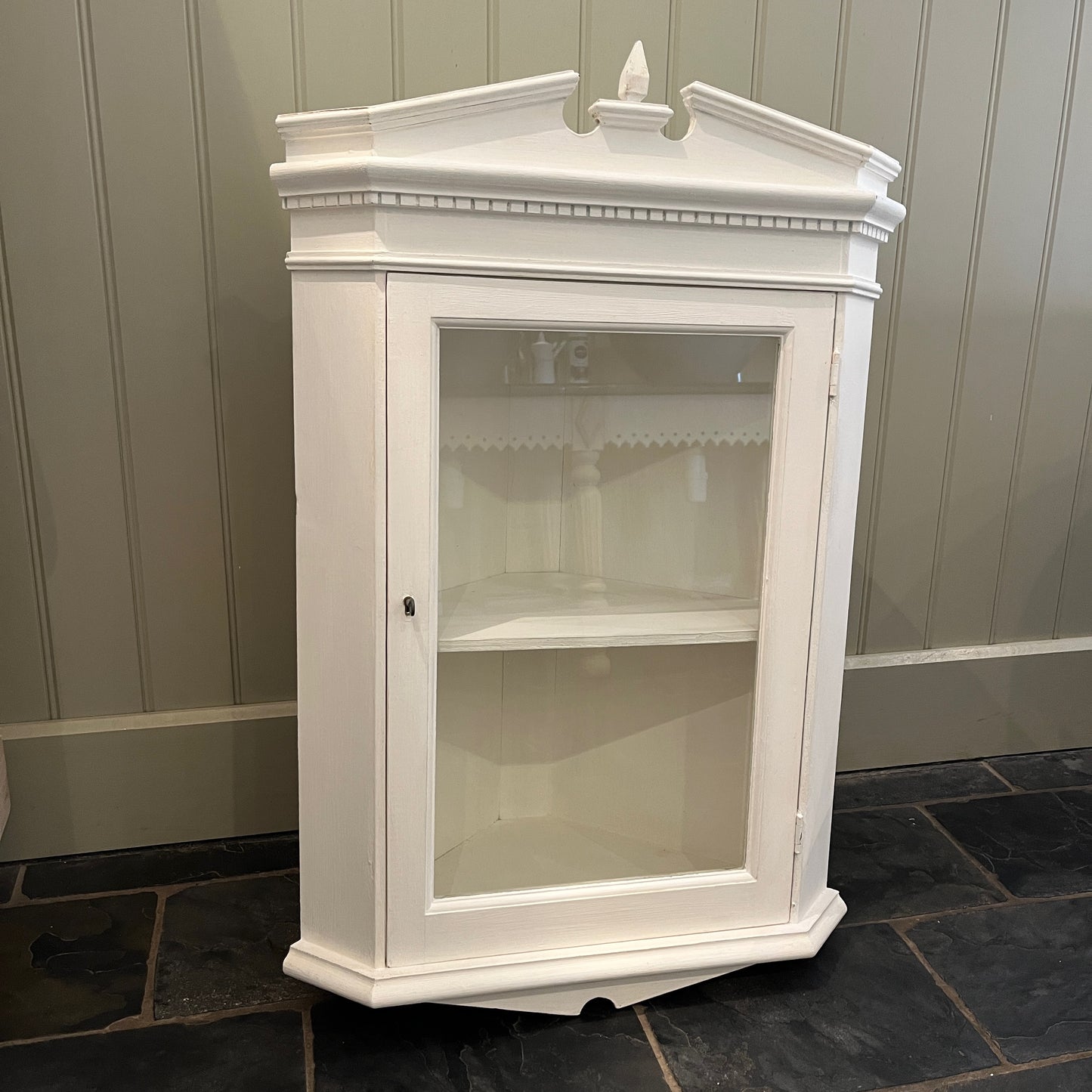A white wooden corner cabinet designed for bathroom use, which fits into a corner, with a single door and interior shelves for practical storage, offering a clean and classic look.