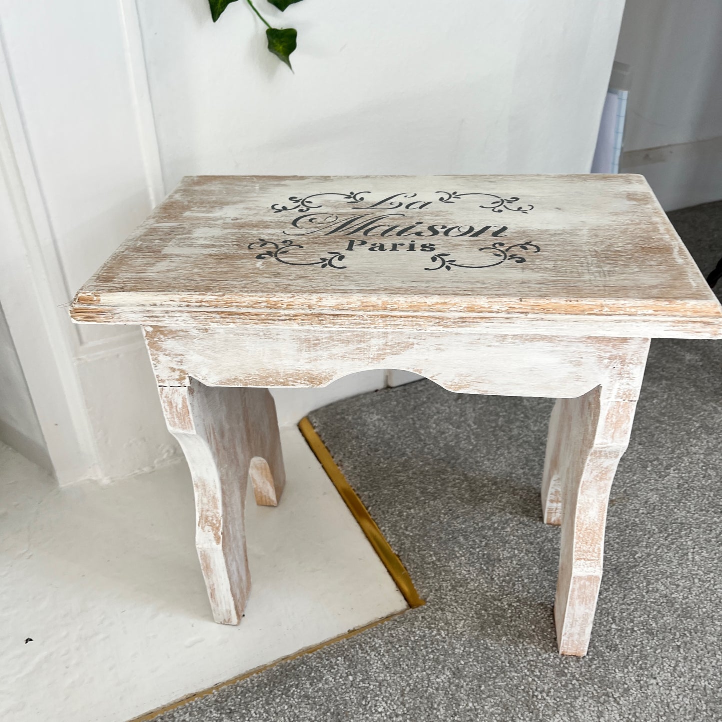 A vintage white wooden stool with a French design stencilled on the top, combining rustic appeal with elegant flair