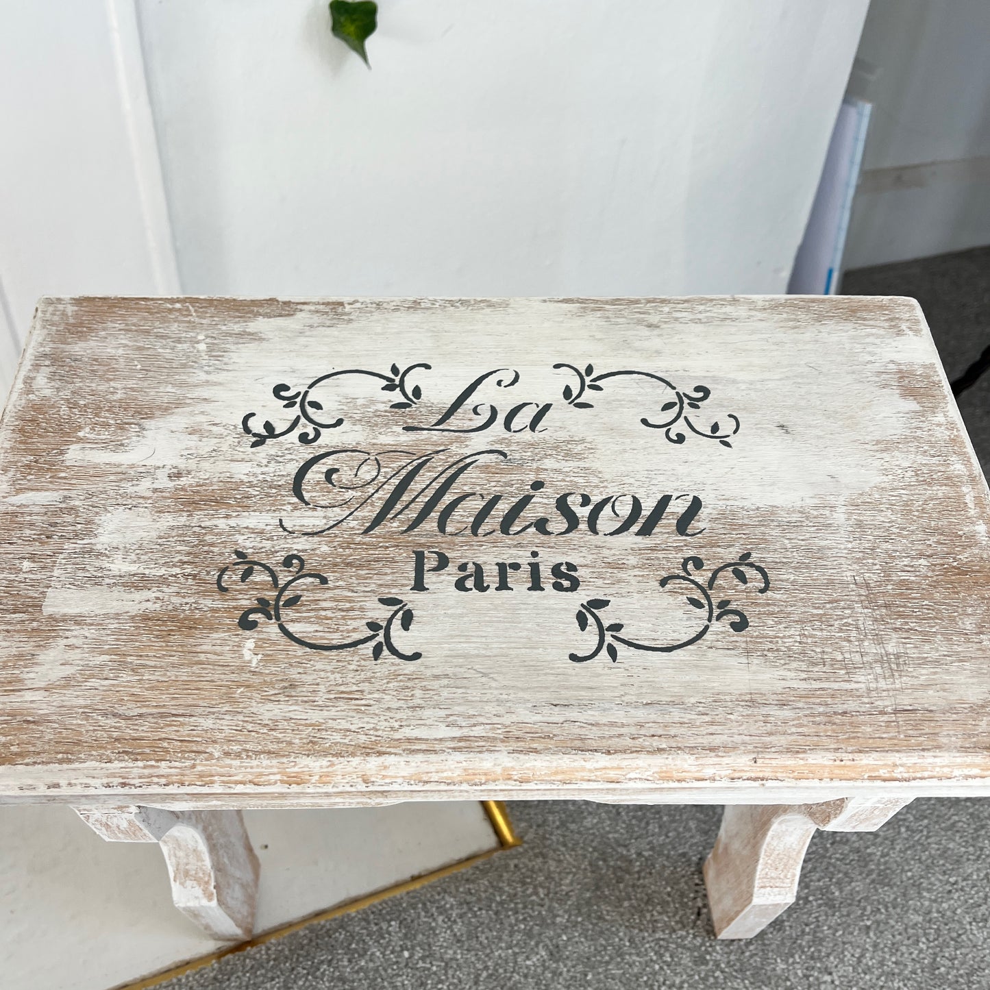 A vintage white wooden stool with a French design stencilled on the top, combining rustic appeal with elegant flair