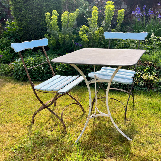 A French vintage bistro set featuring two blue wooden chairs with slightly weathered finishes and curved backs, paired with a square galvanized metal table. The table has a rustic, industrial look with a smooth, silver surface, exuding classic outdoor charm