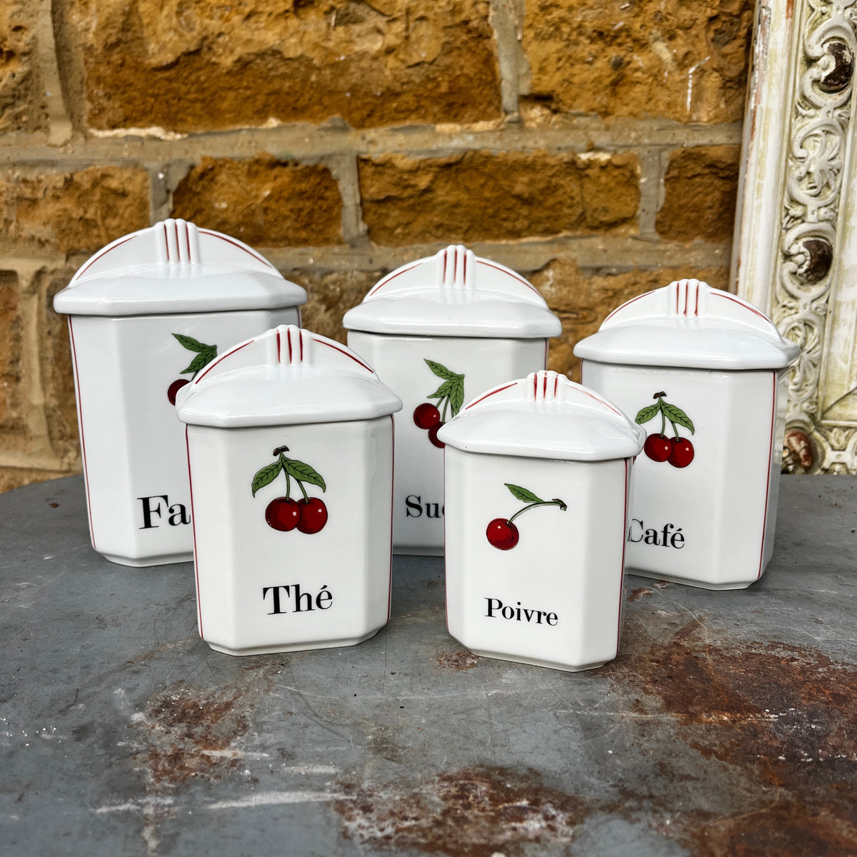 Five white ceramic kitchen canisters with matching lids, decorated with red cherries and black French text (Thé, Poivre, Café, etc.), arranged on a rustic gray stone surface.
