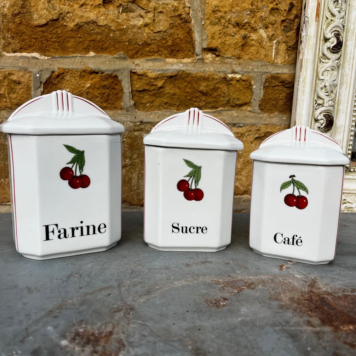 Five white ceramic kitchen canisters with matching lids, decorated with red cherries and black French text (Thé, Poivre, Café, etc.), arranged on a rustic gray stone surface.