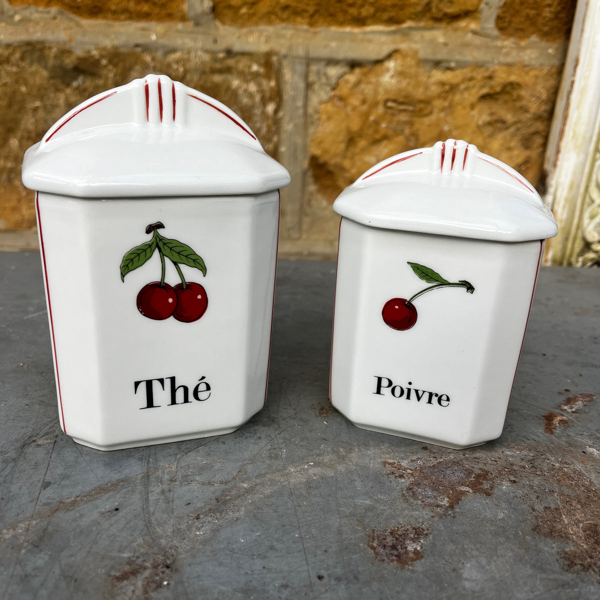 Five white ceramic kitchen canisters with matching lids, decorated with red cherries and black French text (Thé, Poivre, Café, etc.), arranged on a rustic gray stone surface.
