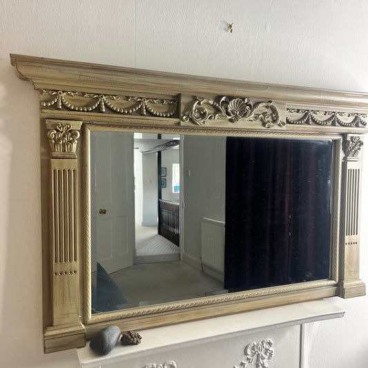 A classic over mantle mirror featuring a grand wooden frame with Romanesque-style columns on either side, adorned with intricate carvings. The beveled mirror surface adds depth and elegance, with a distressed cream finish, exuding luxurious vintage charm