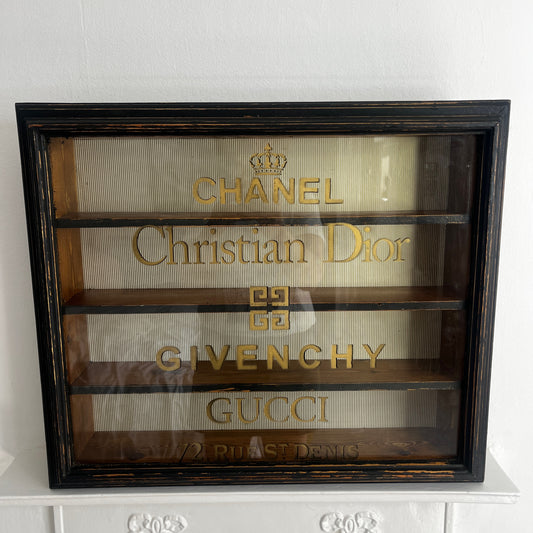 A stylish vintage glass display cabinet with a glass door stencilled with iconic designer names: Chanel, Christian Dior, Givenchy, and Gucci. The cabinet features wooden shelves which could be used to display luxury perfume bottles, fashion books and decorative objects.
