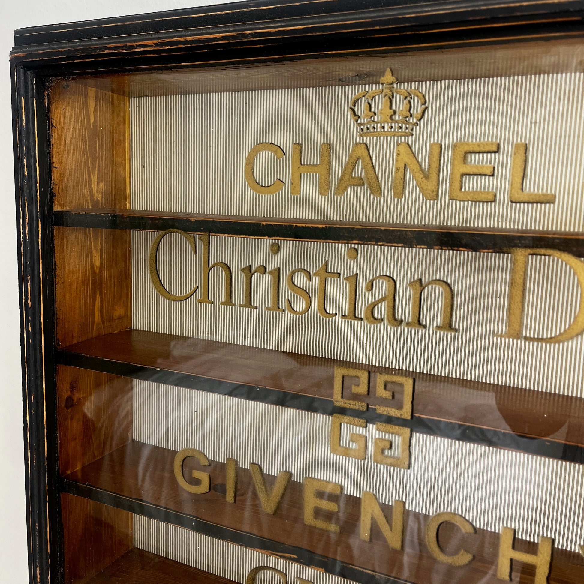 A stylish vintage glass display cabinet with a glass door stencilled with iconic designer names: Chanel, Christian Dior, Givenchy, and Gucci. The cabinet features wooden shelves which could be used to display luxury perfume bottles, fashion books and decorative objects.