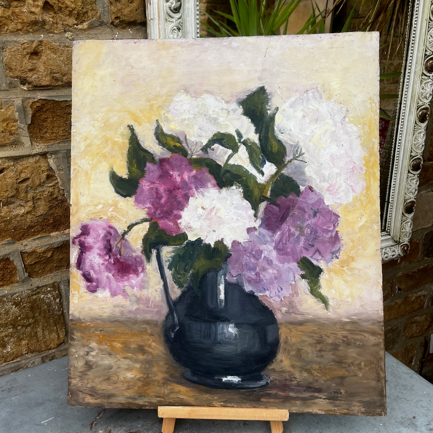 A portrait shape oil painting on board depicting a still life of purple and white flowers in a dark vase, displayed on a small wooden easel.