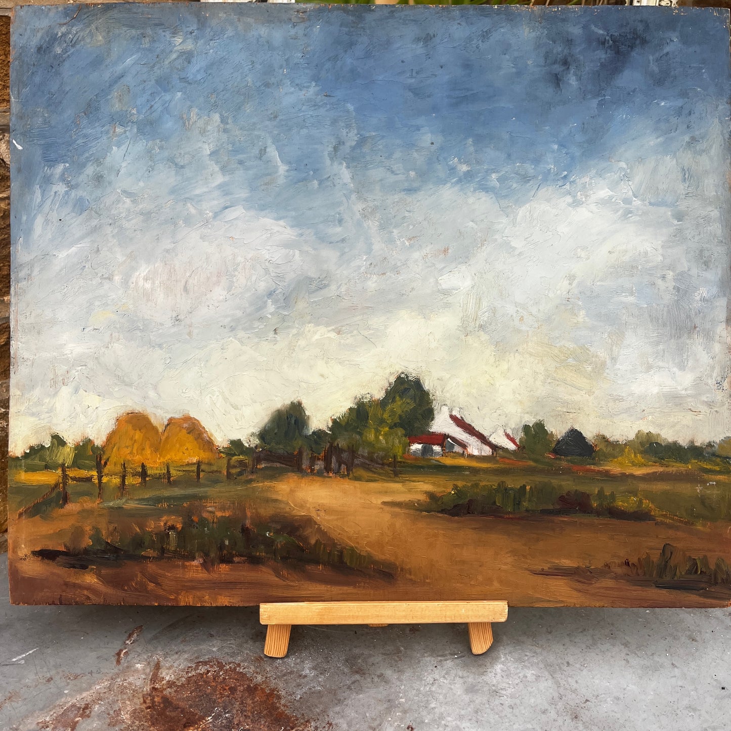 An oil landscape painting on board depicting a rural farm scene with haystacks, trees, a dirt path, and a farmhouse under a cloudy blue sky, displayed on a wooden easel.