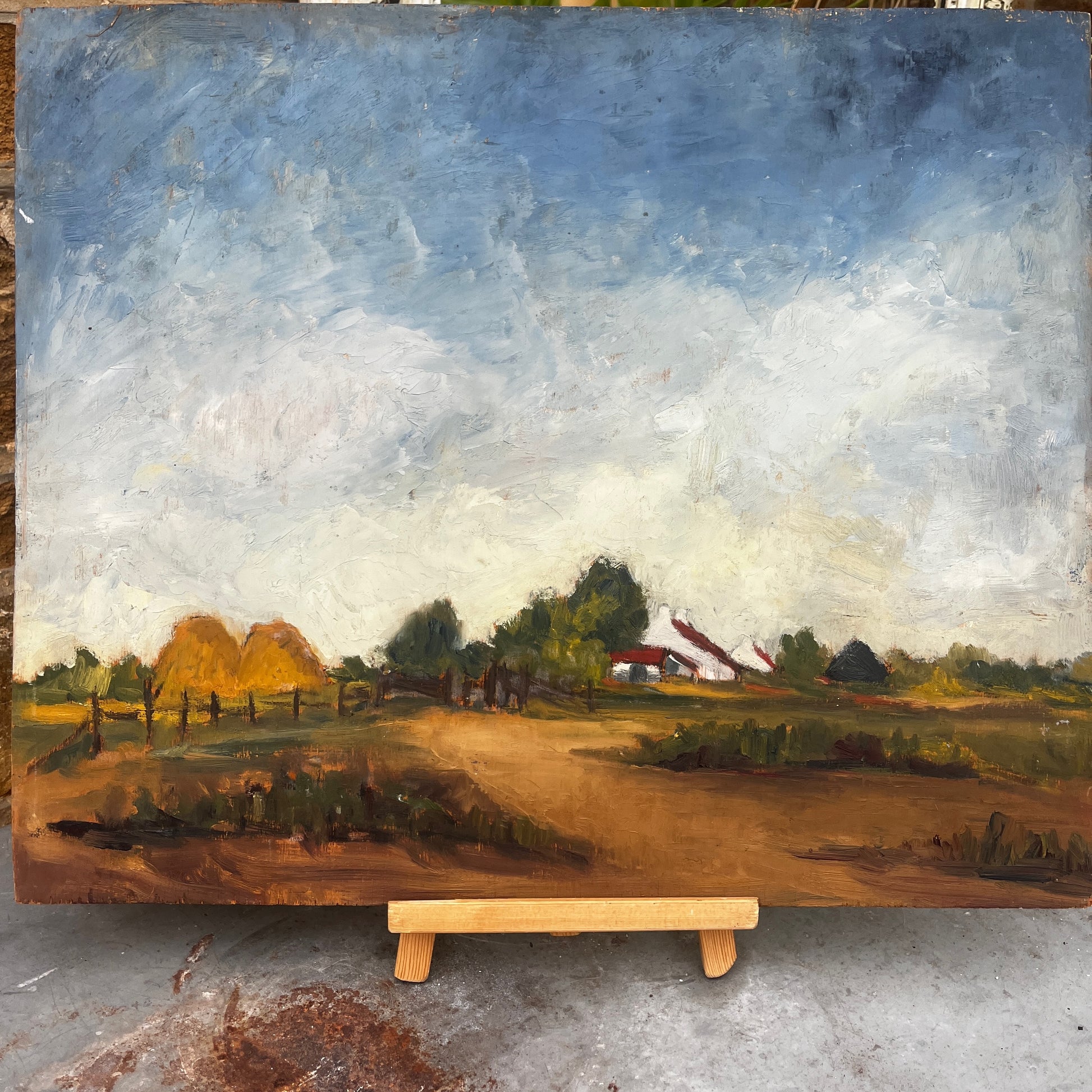 An oil landscape painting on board depicting a rural farm scene with haystacks, trees, a dirt path, and a farmhouse under a cloudy blue sky, displayed on a wooden easel.