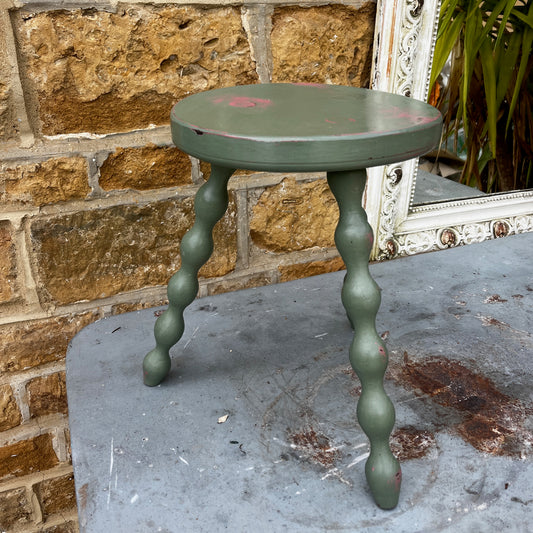 Wooden stool with three turned bobbin-style legs - painted olive green and distressed to show pink coming through.