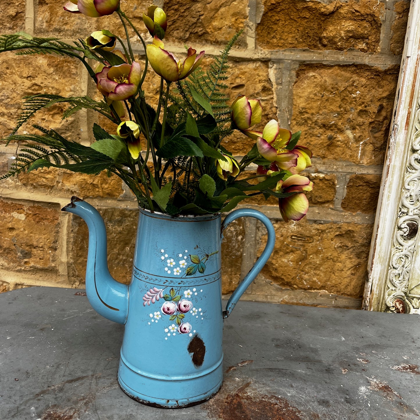 A vintage French enamel jug with a charming floral pattern in soft pink, green, and white tones on a light blue enamel background.