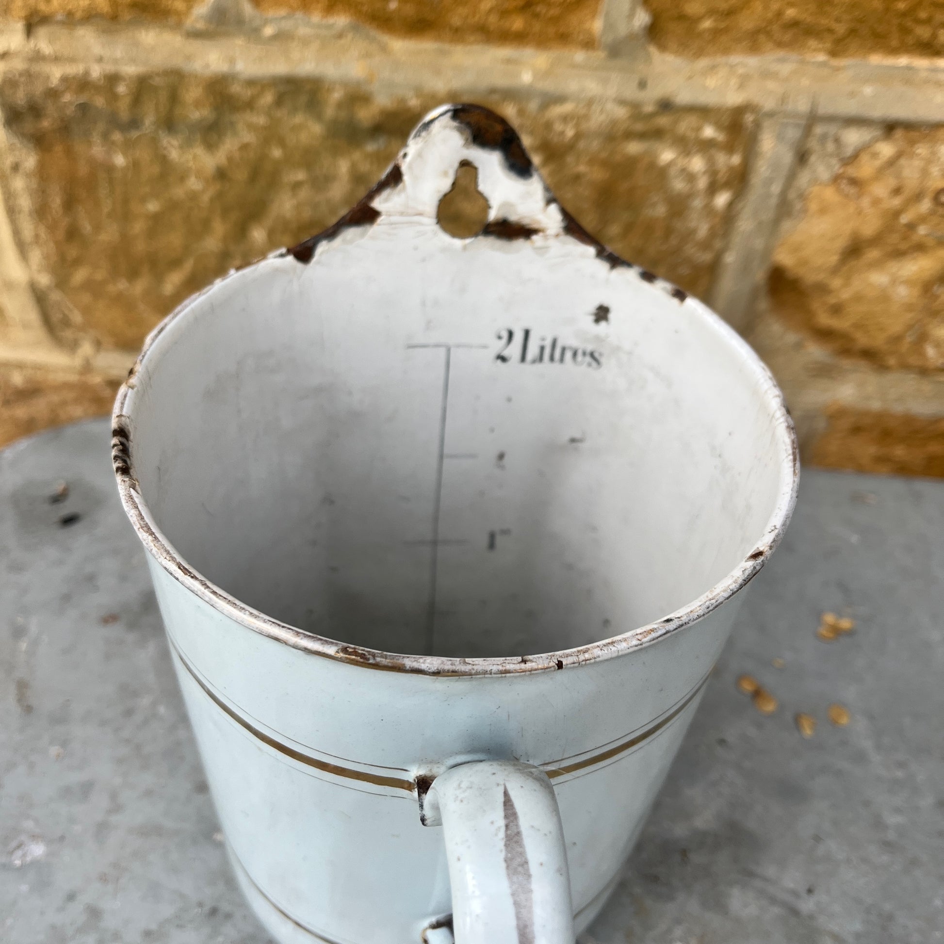 A French irrigation container made of enamel with a white, slightly chipped finish, featuring internal measurement markings etched along the interior wall. The cylindrical container has a sturdy handle and a spout, exuding vintage utility charm