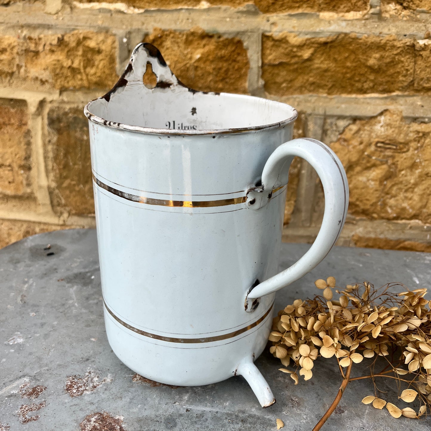 A French irrigation container made of enamel with a white, slightly chipped finish, featuring internal measurement markings etched along the interior wall. The cylindrical container has a sturdy handle and a spout, exuding vintage utility charm