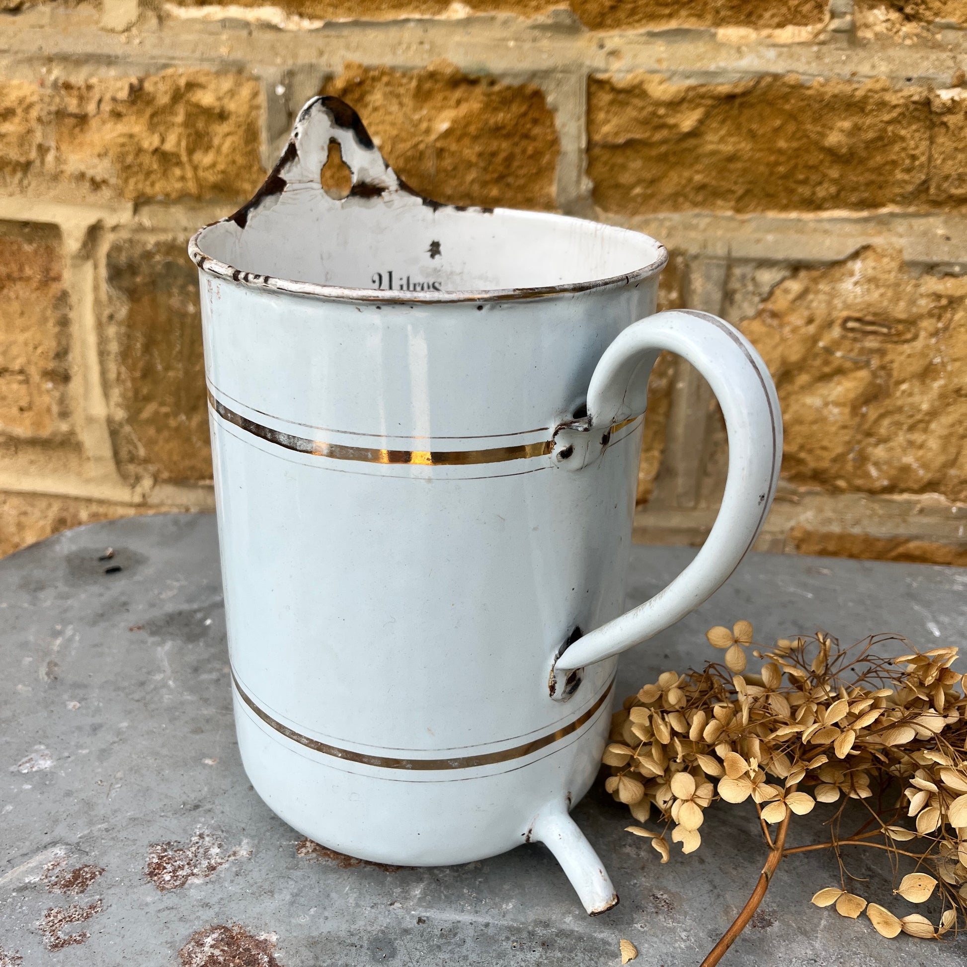 A French irrigation container made of enamel with a white, slightly chipped finish, featuring internal measurement markings etched along the interior wall. The cylindrical container has a sturdy handle and a spout, exuding vintage utility charm