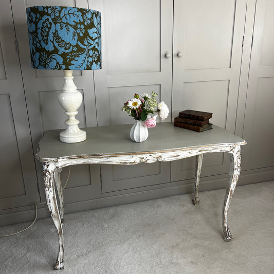 A distressed French-style console table with elegant curved legs. The tabletop is painted a soft gray/green, while the carved wooden frame has a heavily weathered white finish that reveals the wood beneath.