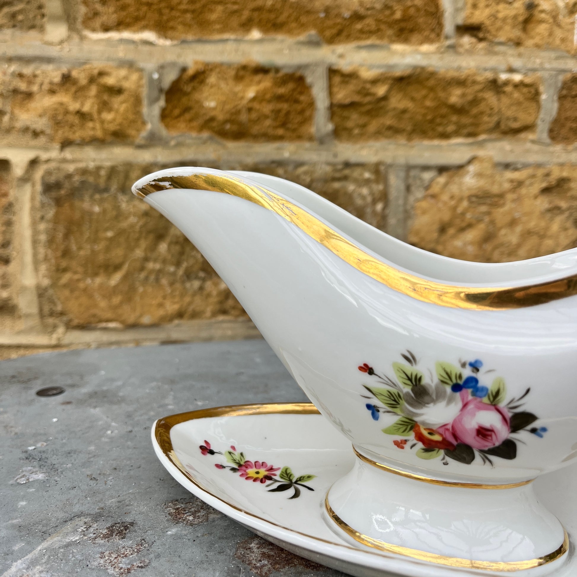 French vintage sauce boat with a fixed saucer, decorated with delicate hand-painted floral motifs and gilded accents along the edges. The piece has an elegant, classic design reflecting refined French tableware style.