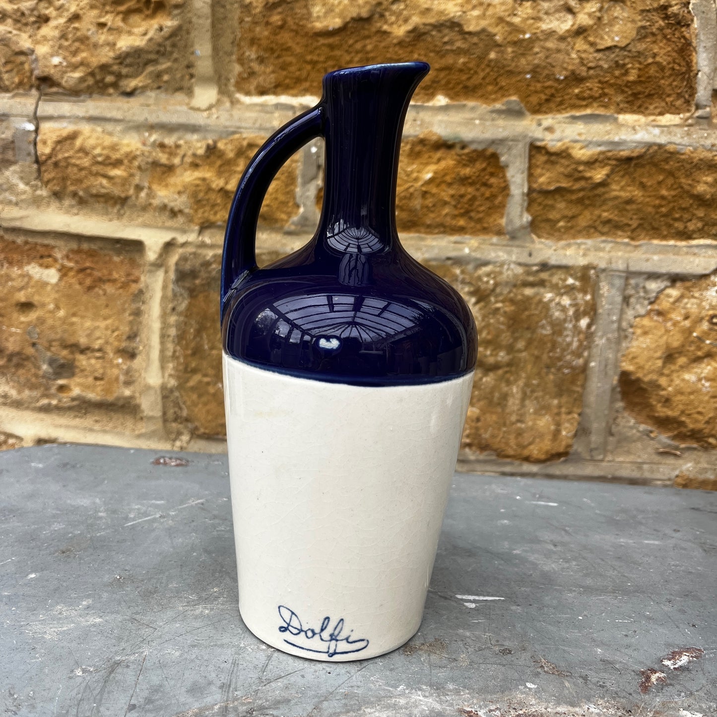 Navy blue and white ceramic jug with 'Dolphi' branding against a stone wall.
