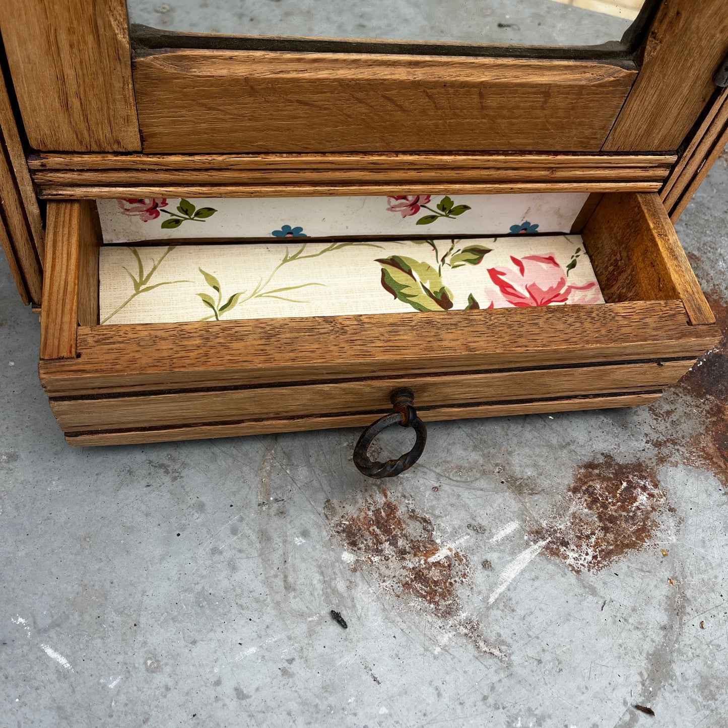 Vintage French wall cupboard with a mirrored door and a single drawer beneath. The interior of the cupboard and the base of the drawer are lined with decorative French paper, adding character and charm to the aged wooden piece.