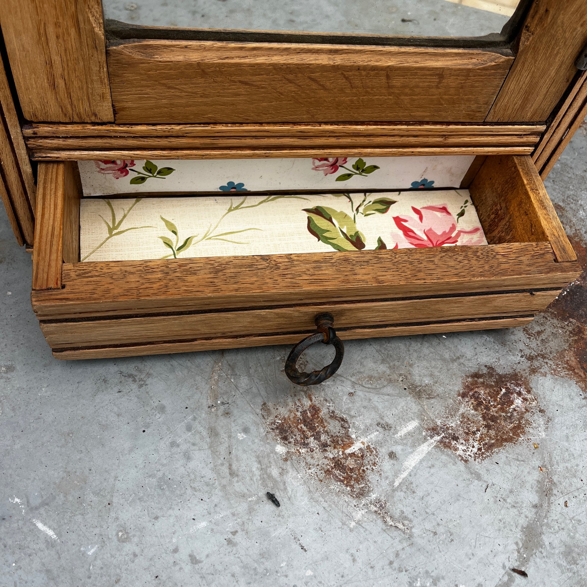 Vintage French wall cupboard with a mirrored door and a single drawer beneath. The interior of the cupboard and the base of the drawer are lined with decorative French paper, adding character and charm to the aged wooden piece.