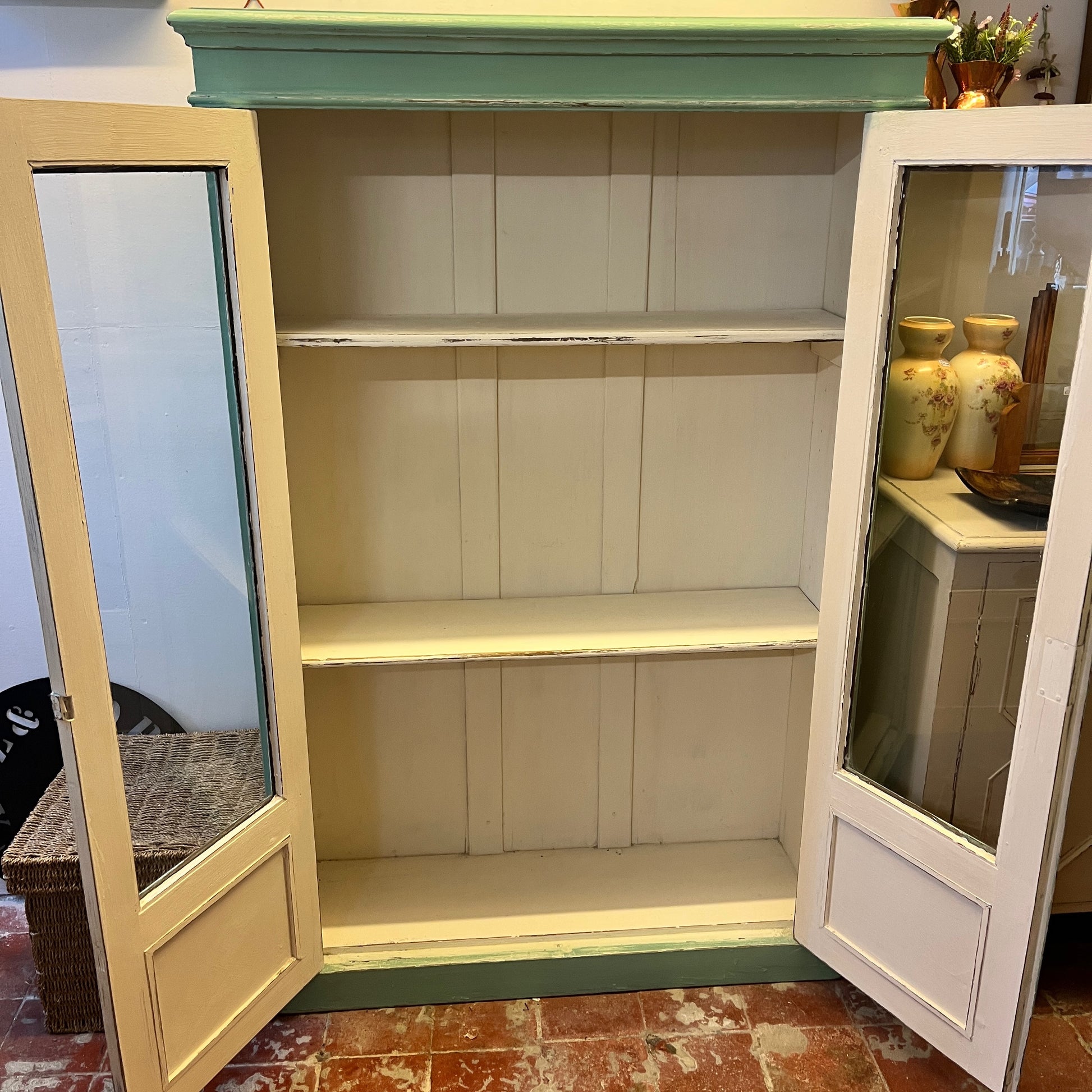 A green vintage cupboard featuring tall panelled glass doors with wooden frames, showcasing a white panelled interior. The cupboard has a classic, slightly distressed finish, highlighting its aged charm. Shelves inside are neatly divided, and the overall design evokes a rustic, farmhouse style.