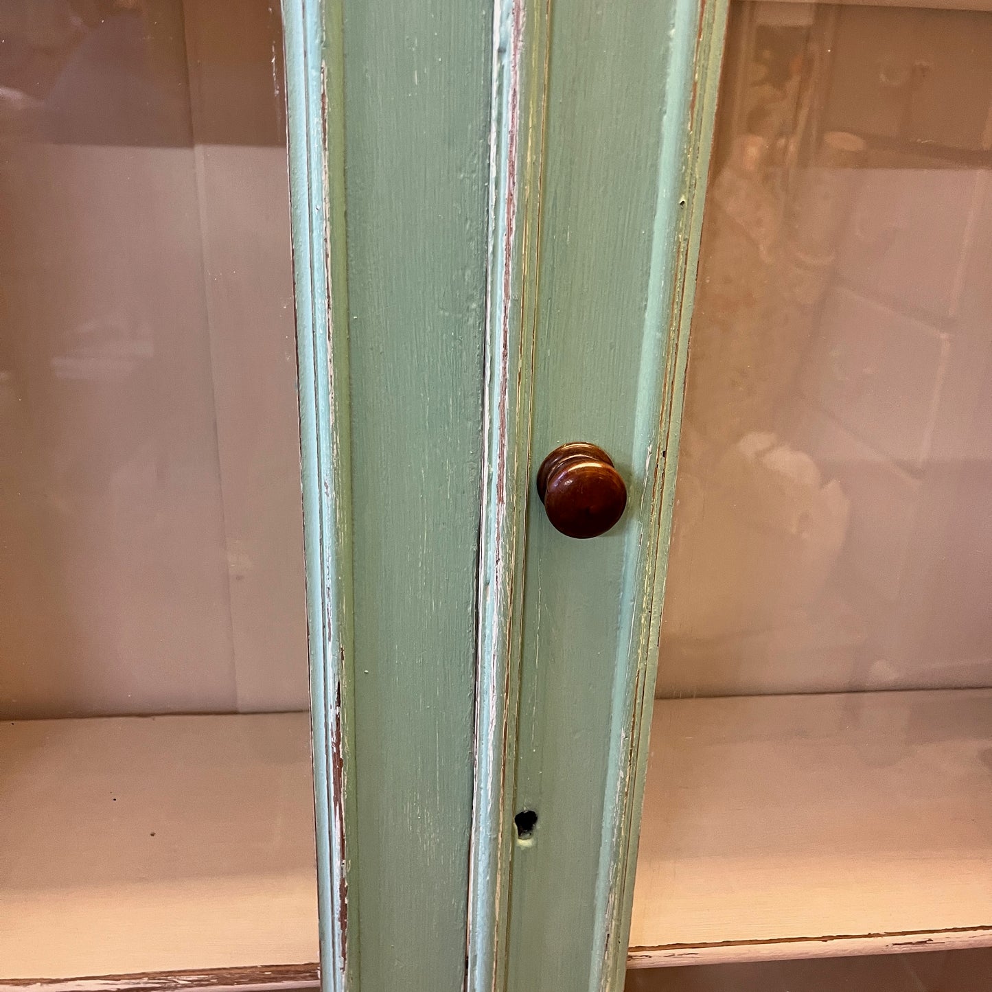 A green vintage cupboard featuring tall panelled glass doors with wooden frames, showcasing a white panelled interior. The cupboard has a classic, slightly distressed finish, highlighting its aged charm. Shelves inside are neatly divided, and the overall design evokes a rustic, farmhouse style.