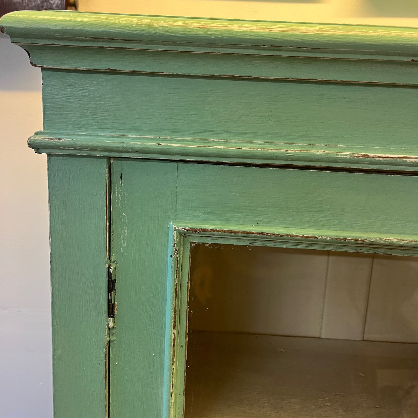 A green vintage cupboard featuring tall panelled glass doors with wooden frames, showcasing a white panelled interior. The cupboard has a classic, slightly distressed finish, highlighting its aged charm. Shelves inside are neatly divided, and the overall design evokes a rustic, farmhouse style.