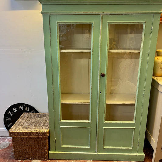 A green vintage cupboard featuring tall panelled glass doors with wooden frames, showcasing a white panelled interior. The cupboard has a classic, slightly distressed finish, highlighting its aged charm. Shelves inside are neatly divided, and the overall design evokes a rustic, farmhouse style.
