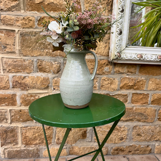 A small vintage garden table crafted from metal with a weathered green finish, featuring a foldable design with hinged legs for easy storage. The table has a simple, circular top with slightly chipped paint, exuding rustic charm