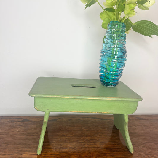 A rustic wooden stool painted in a weathered green finish, showcasing a distressed texture with visible wood grain.