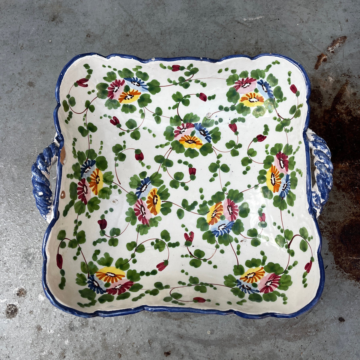 A square ceramic serving dish with scalloped edges and side handles, decorated with a colorful floral pattern on an off-white background.