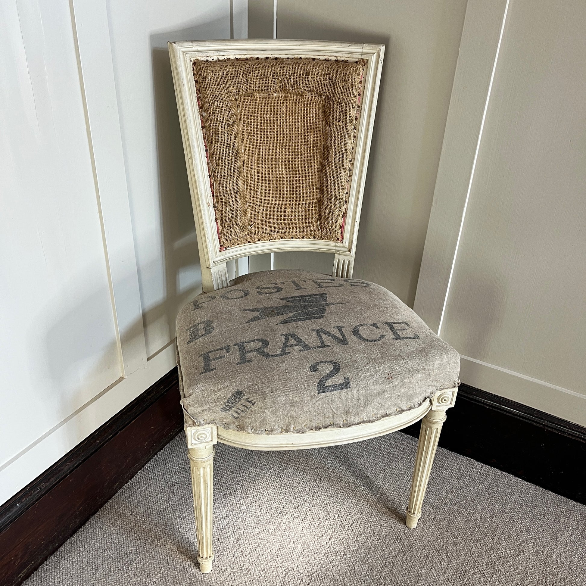 A French chair with a semi-upholstered design, featuring a wooden frame painted in a distressed finish and a back & seat covered in La Poste fabric with its iconic grey and black postal pattern. The chair combines rustic elegance with vintage charm