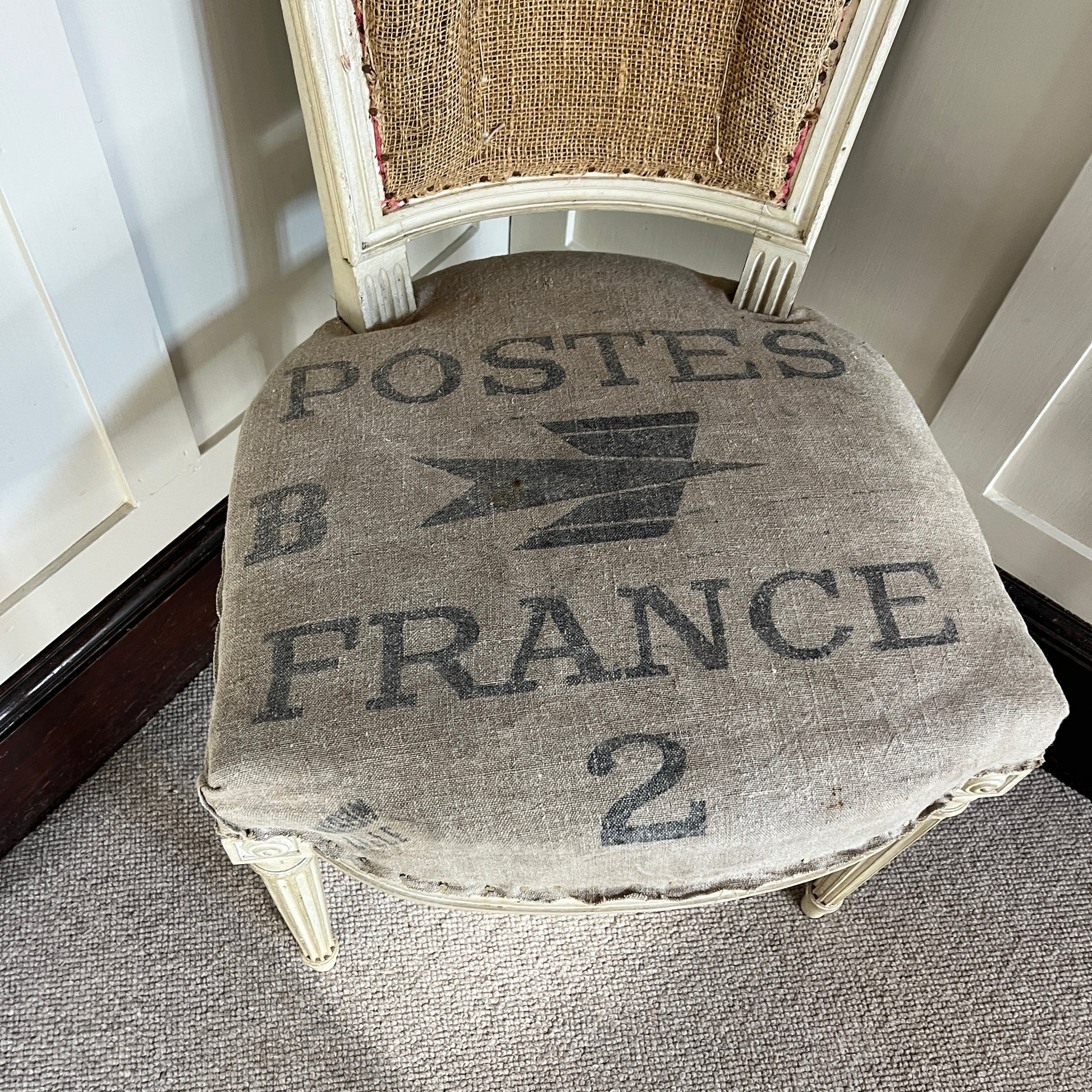A French chair with a semi-upholstered design, featuring a wooden frame painted in a distressed finish and a back & seat covered in La Poste fabric with its iconic grey and black postal pattern. The chair combines rustic elegance with vintage charm