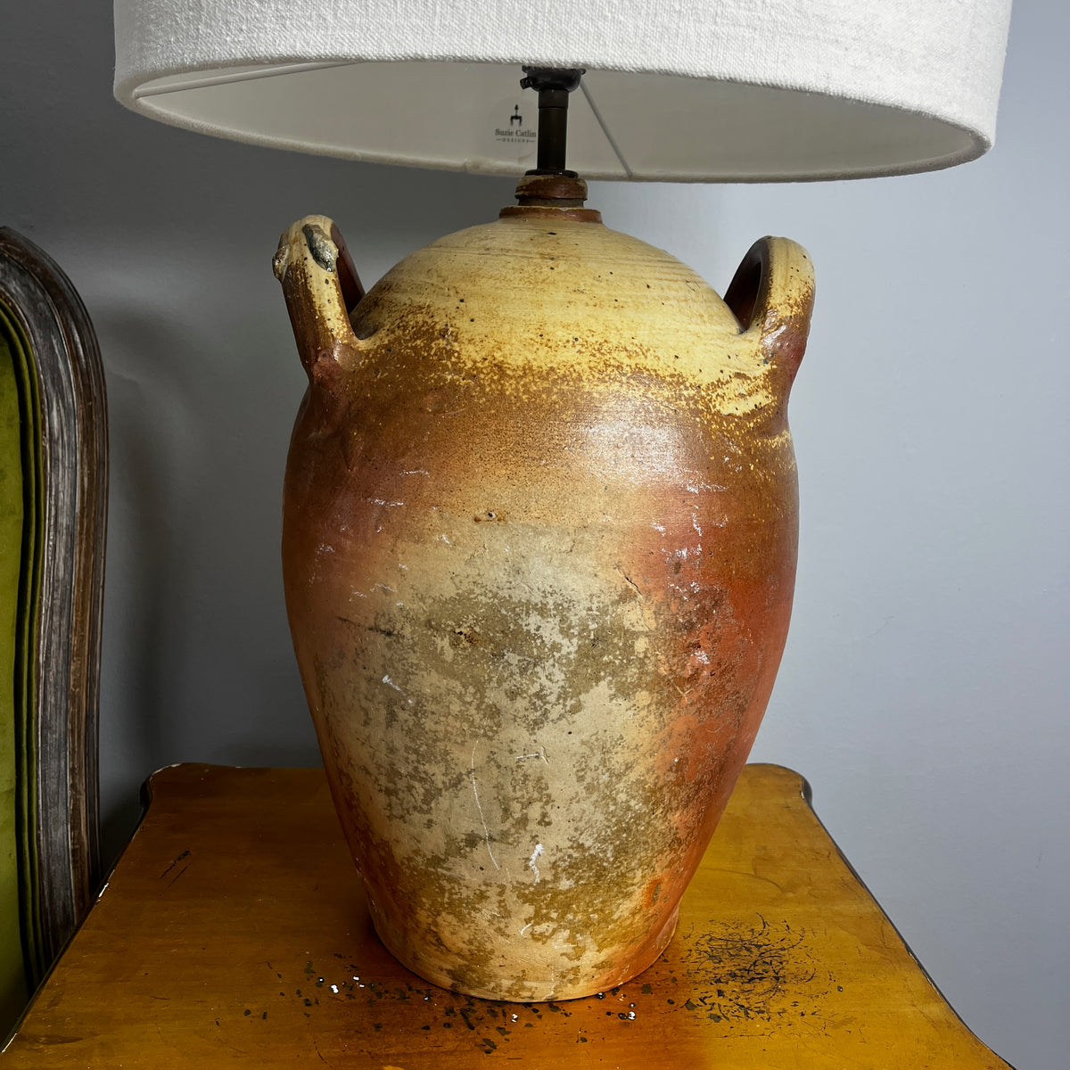 A table lamp with a textured beige linen drum shade and a rustic brown ceramic base with two side handles, shown lit on a wooden bedside table