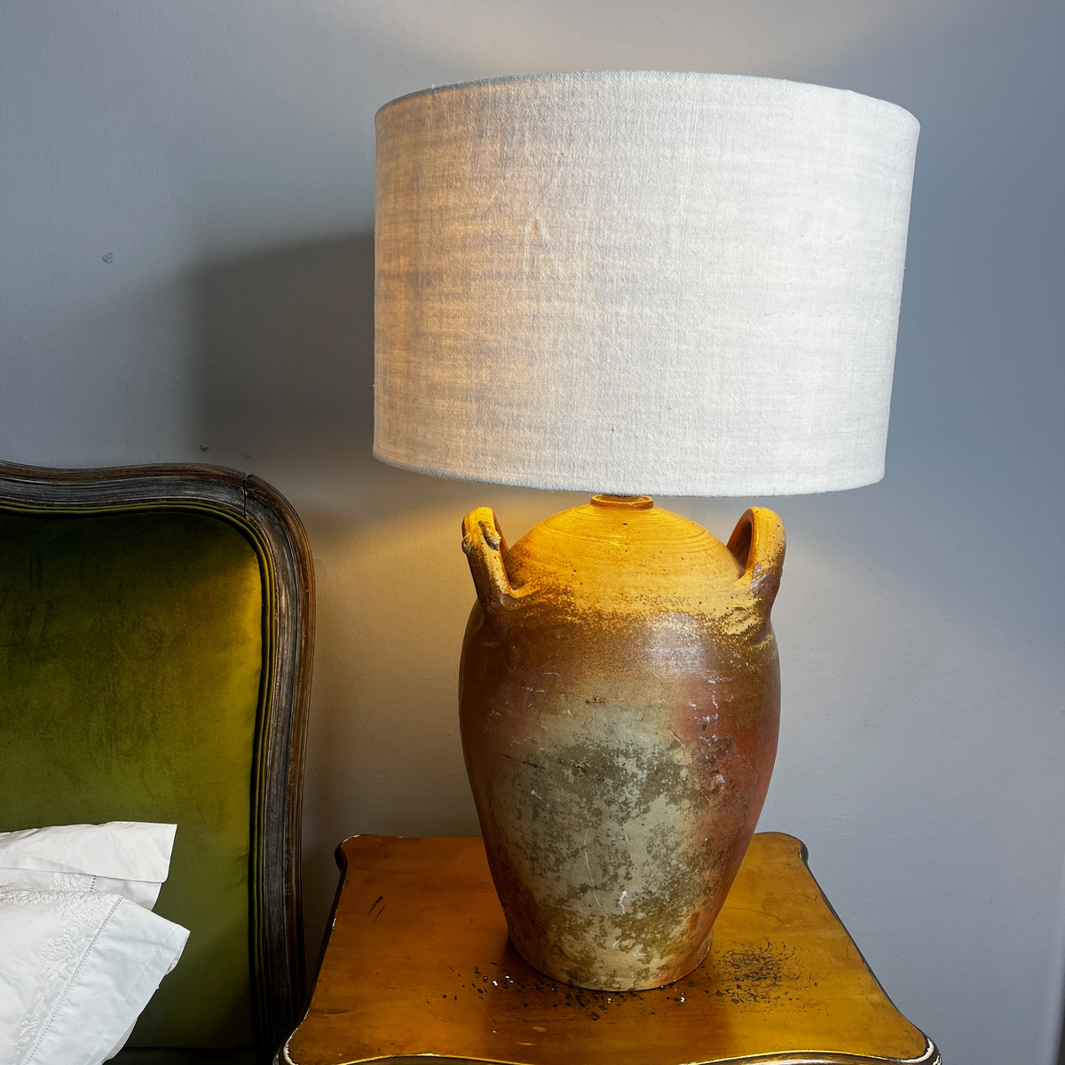 A table lamp with a textured beige linen drum shade and a rustic brown ceramic base with two side handles, shown lit on a wooden bedside table