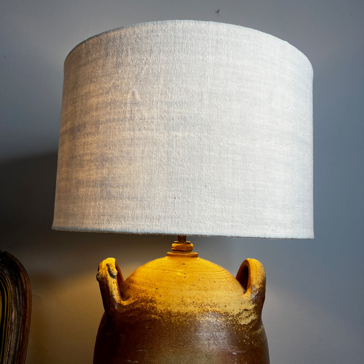 A table lamp with a textured beige linen drum shade and a rustic brown ceramic base with two side handles, shown lit on a wooden bedside table