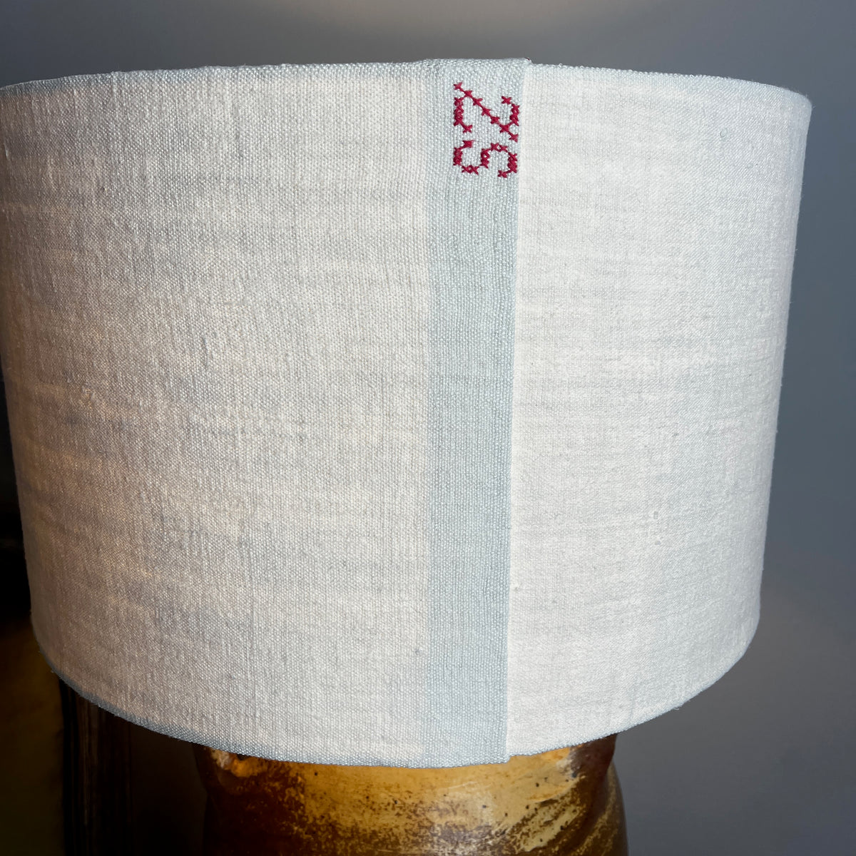 A table lamp with a textured beige linen drum shade and a rustic brown ceramic base with two side handles, shown lit on a wooden bedside table