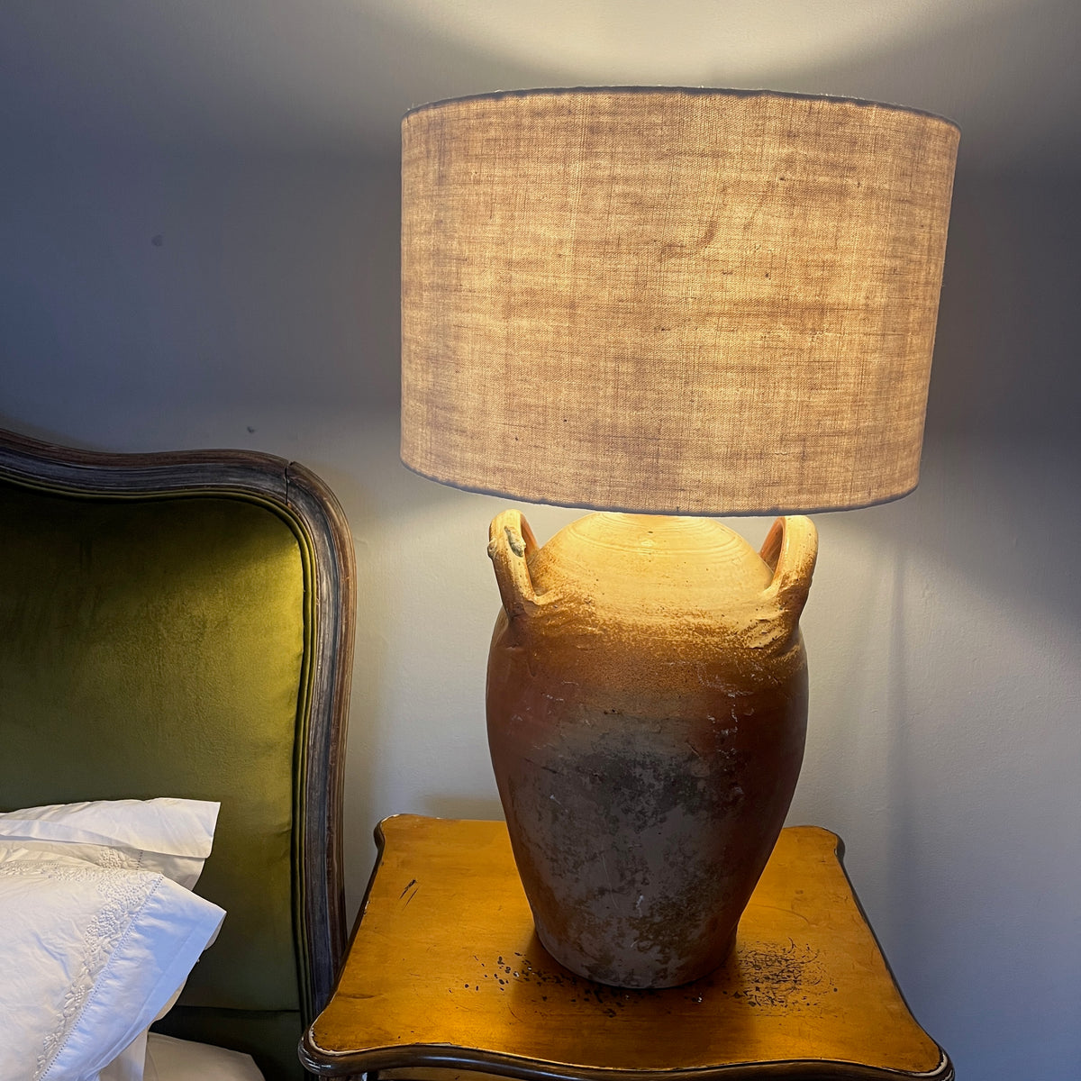 A table lamp with a textured beige linen drum shade and a rustic brown ceramic base with two side handles, shown lit on a wooden bedside table