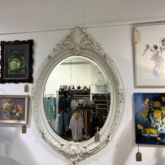 A large oval vintage mirror with a bevelled glass edge, framed by an ornate white wooden frame featuring intricate floral carvings