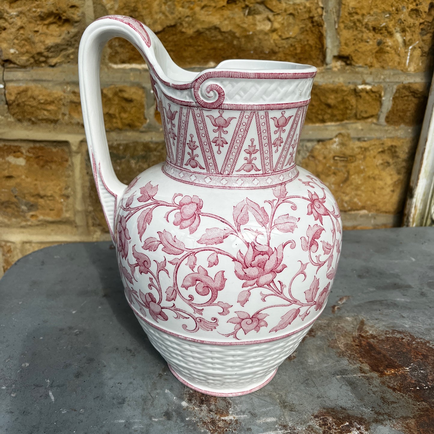 An antique French Sarreguemines ironstone pitcher featuring a classic pink transferware floral pattern on a creamy white background. The generously sized pitcher has a curved handle, gently flared rim, and elegant rounded form, with subtle signs of age and wear that enhance its vintage character.