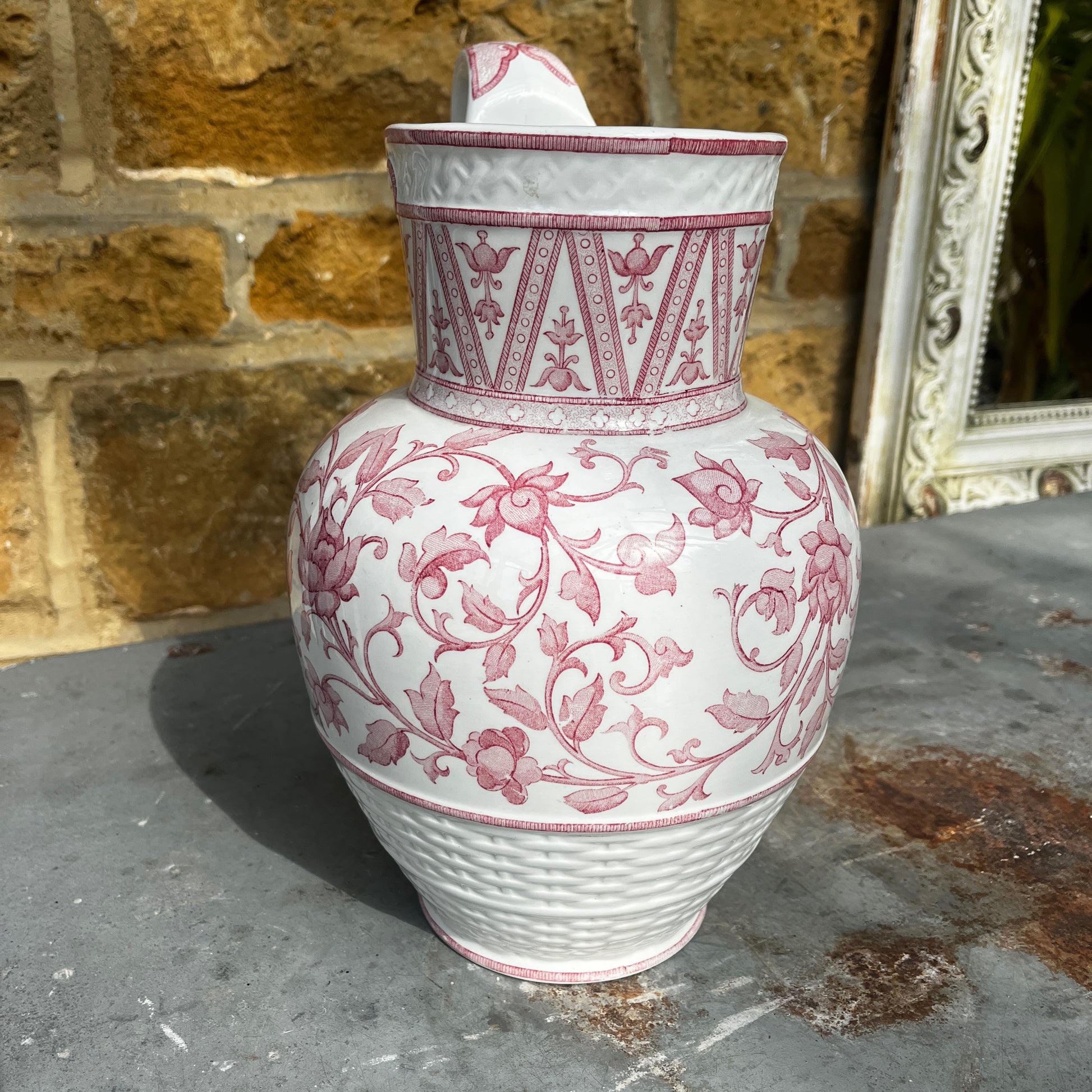 An antique French Sarreguemines ironstone pitcher featuring a classic pink transferware floral pattern on a creamy white background. The generously sized pitcher has a curved handle, gently flared rim, and elegant rounded form, with subtle signs of age and wear that enhance its vintage character.