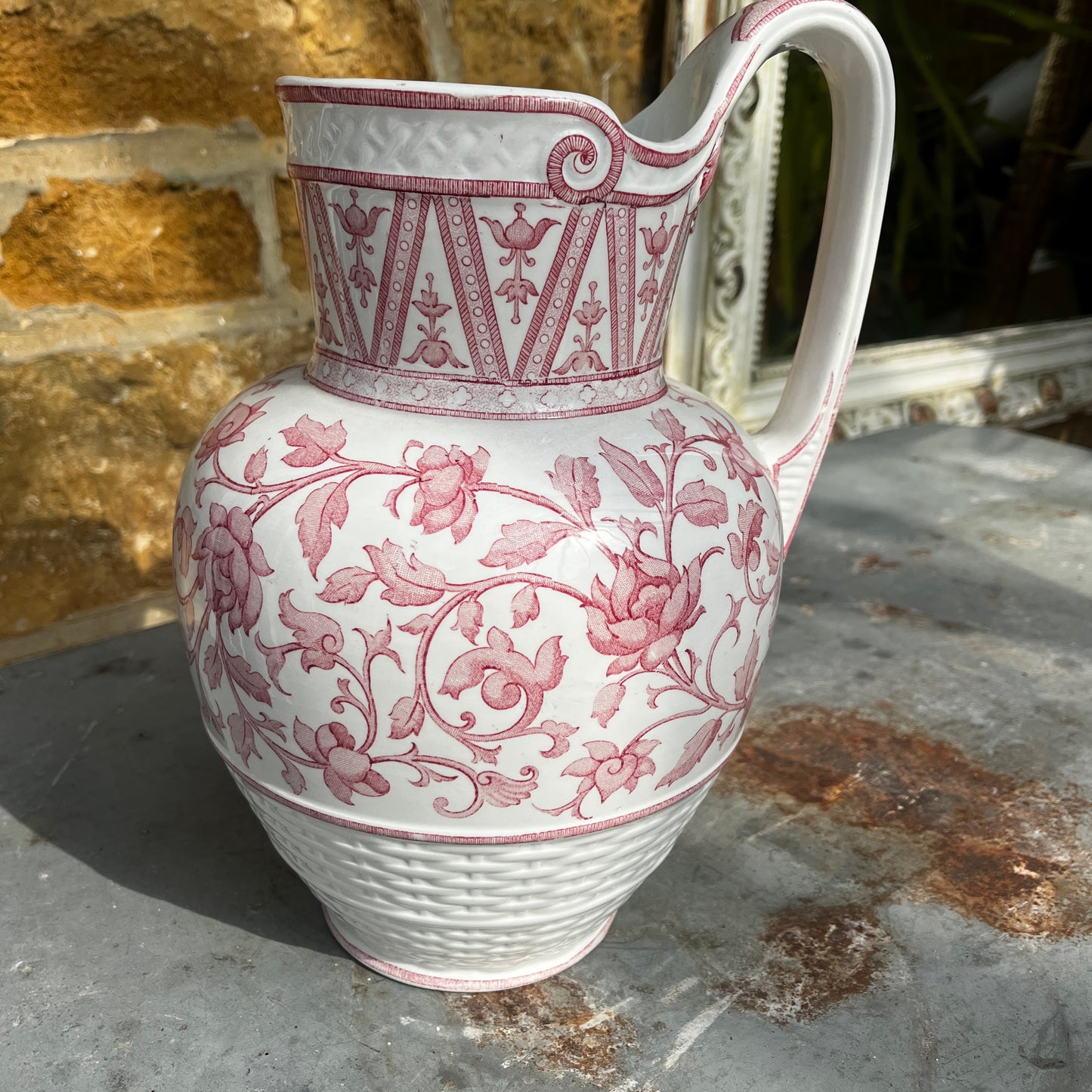 An antique French Sarreguemines ironstone pitcher featuring a classic pink transferware floral pattern on a creamy white background. The generously sized pitcher has a curved handle, gently flared rim, and elegant rounded form, with subtle signs of age and wear that enhance its vintage character.