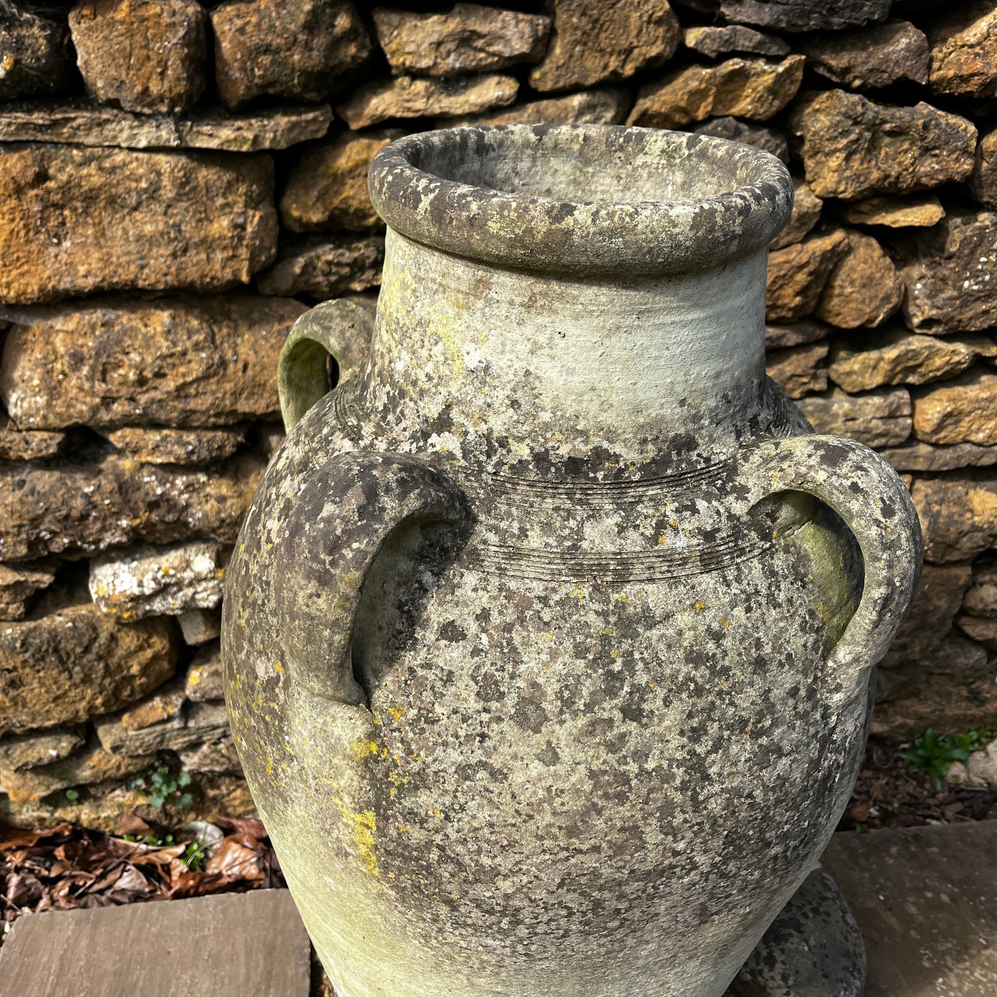 Large rustic garden urn with an aged weathered surface and traces of green lichen, featuring a classic decorative shape and timeworn outdoor character.
