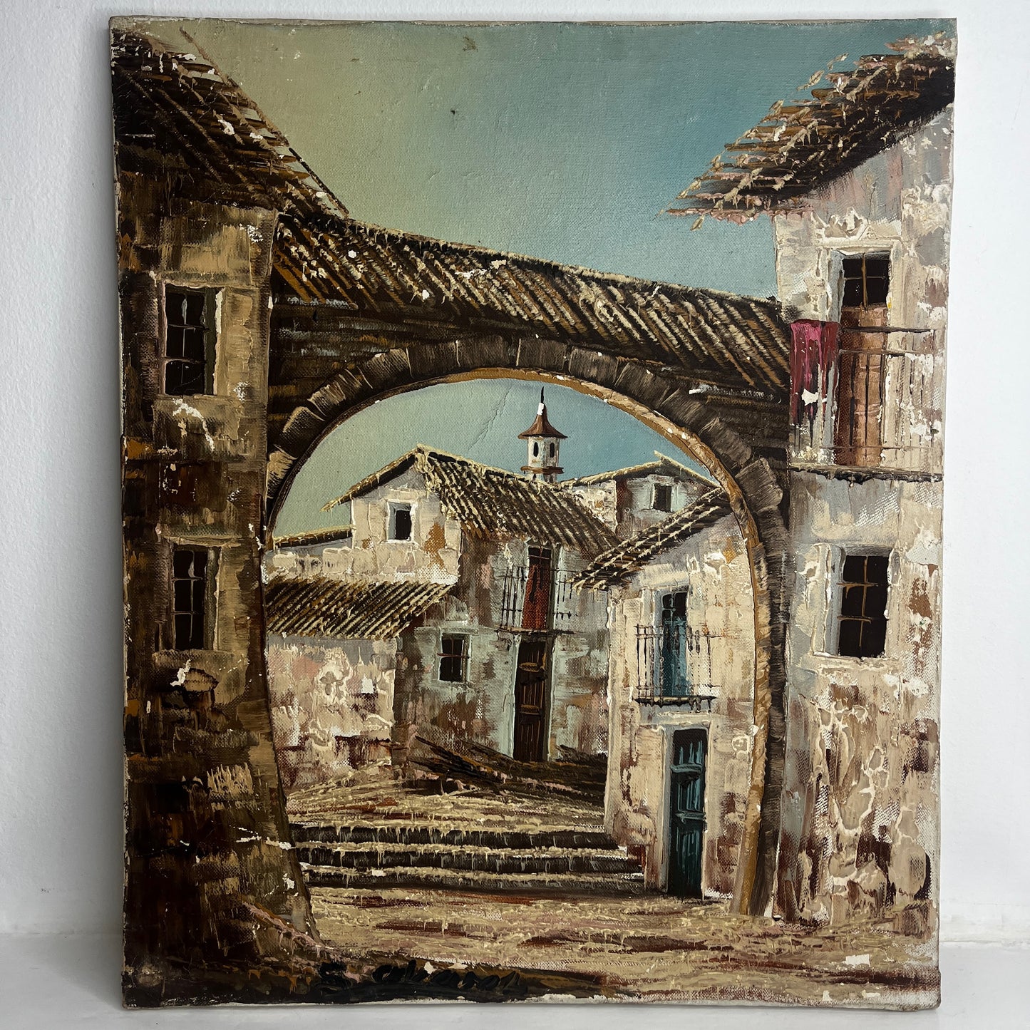 An oil painting depicting a rustic French village scene viewed through a large stone archway, with weathered buildings, tiled roofs, and a pale blue sky.