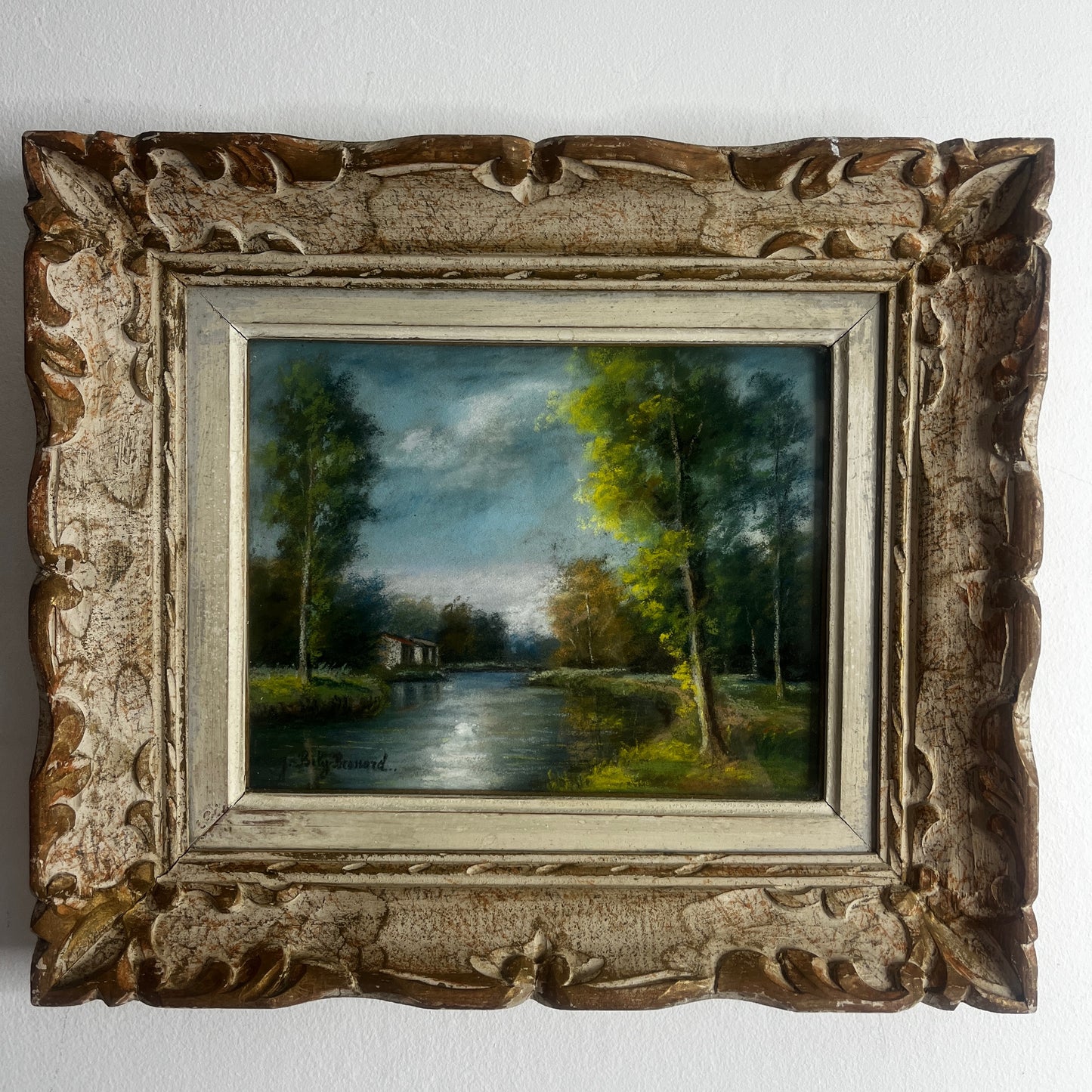 A vintage oil painting of a tranquil wooded landscape featuring a calm river or lake reflecting the sky, with trees lining the water and a small building nestled among the trees in the distance.  Montparnasse Frame.